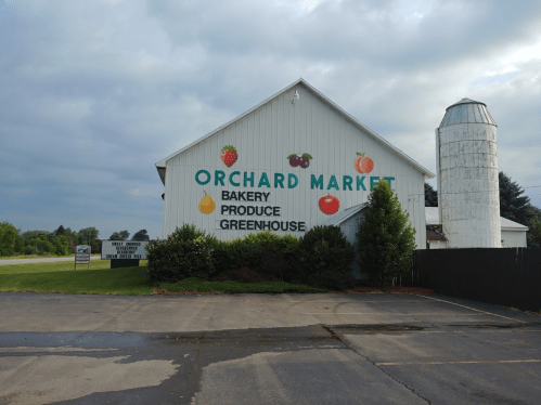A white barn-style building with "Orchard Market" signage, featuring a silo and cloudy sky in the background.