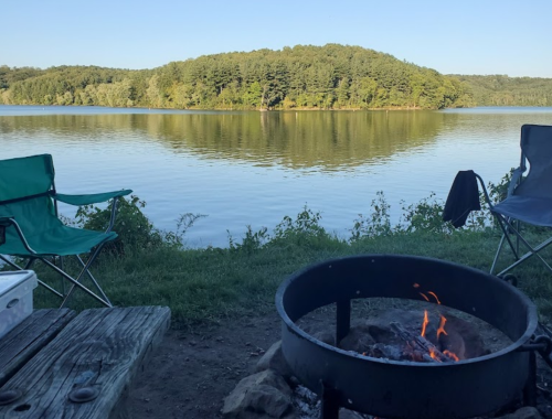 A serene lakeside view with a campfire, two chairs, and an island in the distance surrounded by trees.