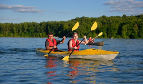 Two people in yellow kayaks paddle on a calm lake, surrounded by trees and blue skies.