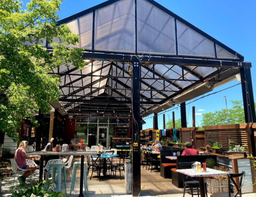 A bright outdoor dining area with tables under a large, open structure surrounded by greenery.
