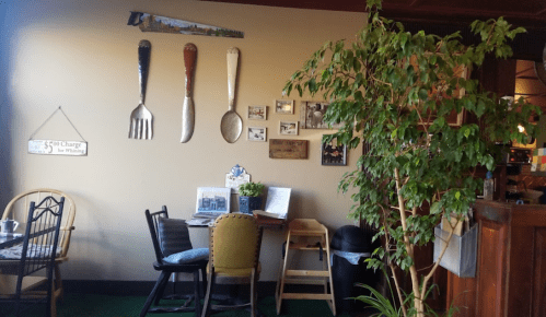 Cozy café corner with large utensils on the wall, a small table, chairs, and a potted plant.