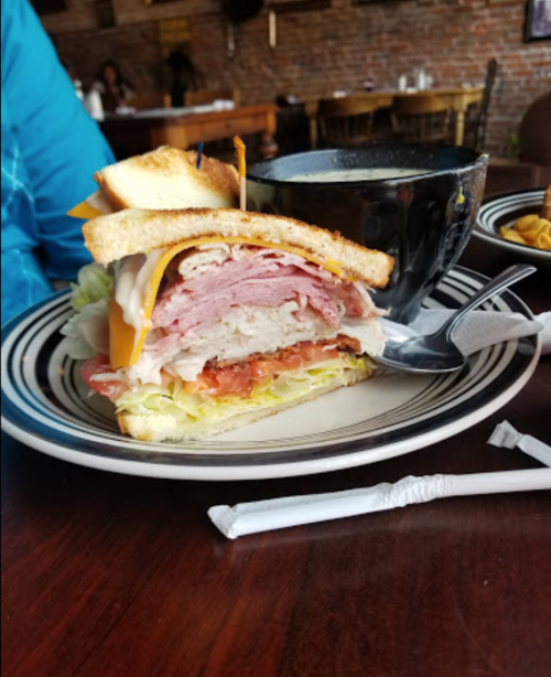 A stacked sandwich with turkey, ham, cheese, lettuce, and tomato, served with a bowl of soup on the side.