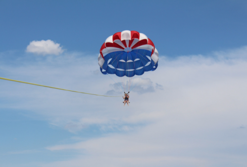 Two people parasailing under a colorful red, white, and blue parachute against a clear blue sky.