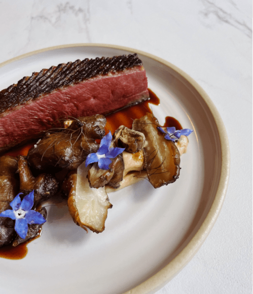 A plate featuring seared steak, sautéed mushrooms, and edible flowers on a light background.