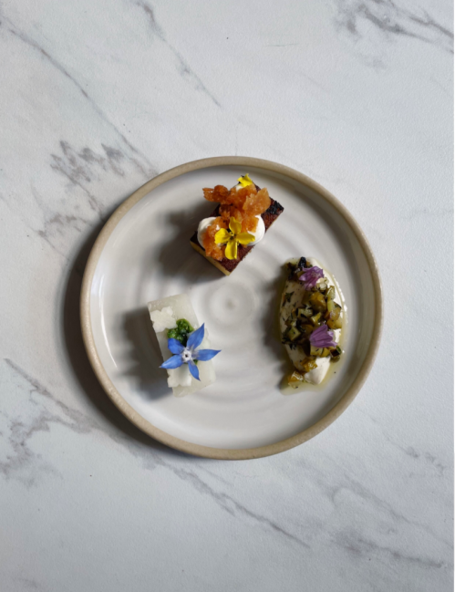 A white plate with three decorative bite-sized dishes, each topped with colorful edible flowers.