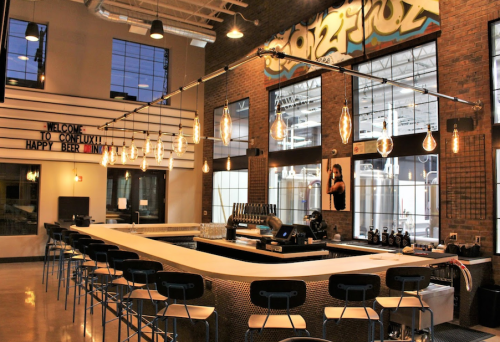 Modern bar interior with a long counter, industrial decor, and large windows, featuring hanging lights and a welcoming sign.
