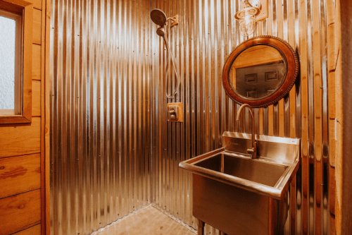 A rustic bathroom with corrugated metal walls, a vintage mirror, a shower, and a stainless steel sink.