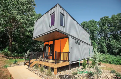 A modern two-story house made from shipping containers, featuring colorful accents and surrounded by greenery.