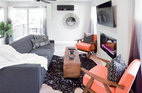 Modern living room with a gray sofa, orange chairs, wooden coffee table, and an electric fireplace. Bright and inviting space.