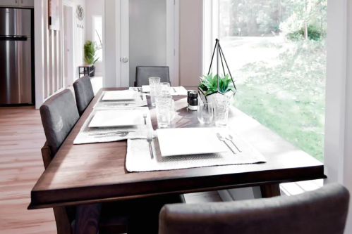 A modern dining table set for four, featuring plates, glasses, and a small plant, with a view of greenery outside.
