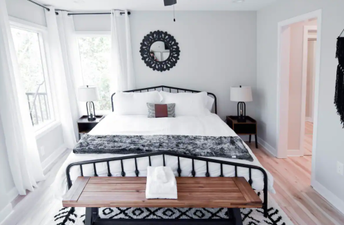Bright, modern bedroom with a large bed, decorative mirror, and wooden bench, featuring white curtains and a cozy rug.