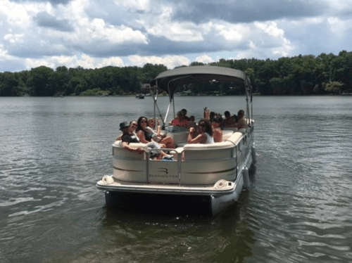 A group of people enjoying a sunny day on a pontoon boat on a calm lake surrounded by trees.