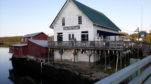 A white country store with a green roof, overlooking a calm waterway, surrounded by red buildings and a clear sky.