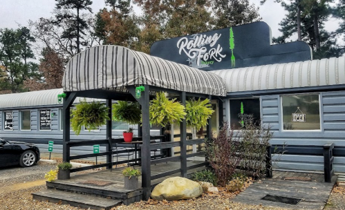 Exterior of Rolling Fork restaurant with a striped awning, potted plants, and a gravel parking area surrounded by trees.