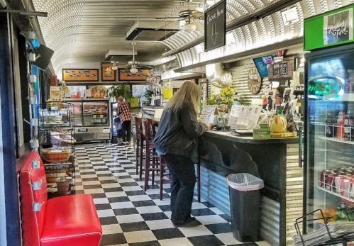 A cozy diner interior with checkered floors, a counter, and patrons enjoying their meals. Brightly lit and inviting atmosphere.