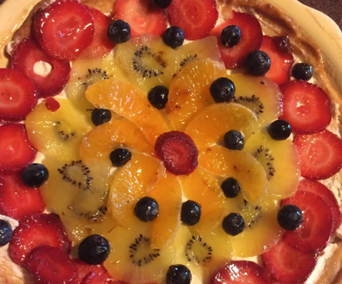 A colorful fruit tart topped with strawberries, kiwi, oranges, and blueberries arranged in a floral pattern.