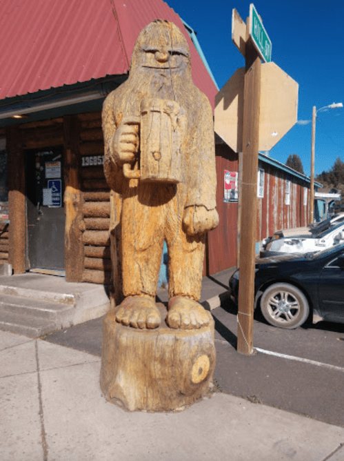 A large wooden statue of a Sasquatch holding a beer mug, standing on a log near a roadside.