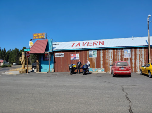 A rustic tavern with a blue roof and red signage, featuring parked motorcycles and a car outside on a sunny day.