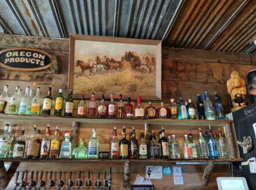 A rustic bar shelf displaying various bottles of liquor, with a vintage horse-drawn wagon painting above.