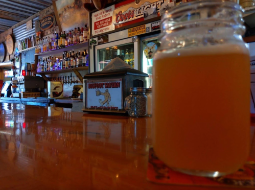 A close-up of a glass of beer on a bar counter, with shelves of liquor and a sign in the background.