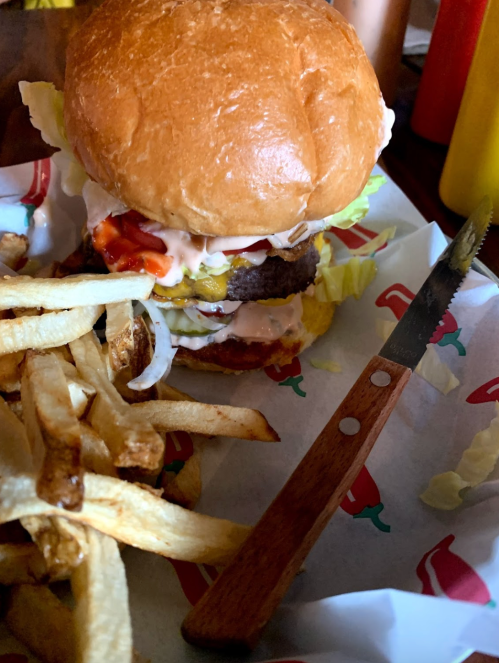 A close-up of a burger with lettuce, pickles, and sauces, alongside a serving of fries and a knife on a patterned paper.
