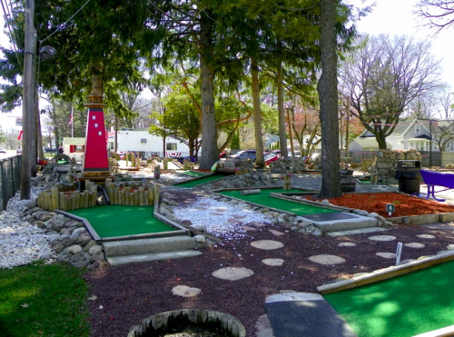 A mini-golf course with green turf, stone pathways, and trees, set in a sunny outdoor environment.