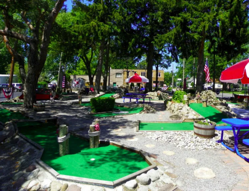 A mini-golf course with green turf, trees, and colorful umbrellas, surrounded by rocks and benches.