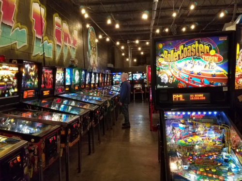 A dimly lit arcade filled with pinball machines, featuring a player focused on a game in the background.