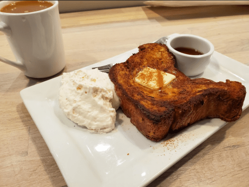 A plate with French toast topped with butter, a side of whipped cream, and a small cup of syrup, with coffee in the background.