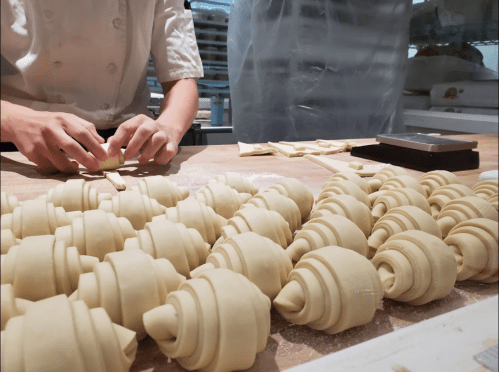 A baker shapes croissant dough on a wooden surface, with neatly arranged uncooked croissants in the foreground.