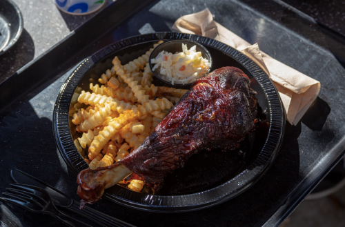 A black plate with a grilled turkey leg, crinkle-cut fries, and a small cup of coleslaw on a tray.