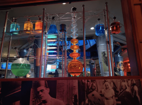 Colorful laboratory glassware filled with liquids, displayed on a metal rack in a vibrant, illuminated setting.