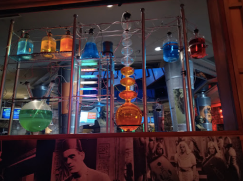 Colorful laboratory glassware filled with liquids, displayed on a metal rack in a vibrant, illuminated setting.