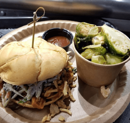 A sandwich with pulled meat and slaw, served with a side of Brussels sprouts and a small cup of sauce.