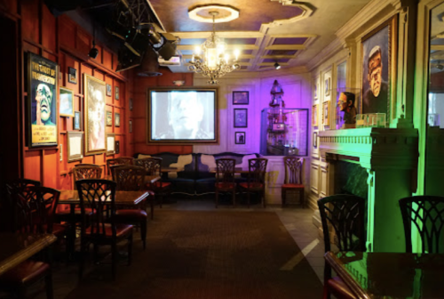 A cozy bar with red walls, vintage decor, and a projector screen displaying a film, illuminated by colorful lights.