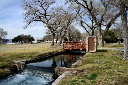 A serene landscape featuring a small stream, a wooden bridge, and trees, with a grassy area in the background.