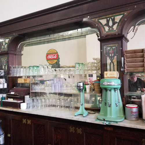 Vintage soda fountain counter with glassware, a green mixer, and a Coca-Cola sign in a nostalgic setting.