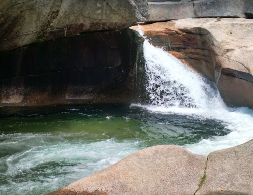 A small waterfall cascades over smooth rocks into a clear, green pool below.