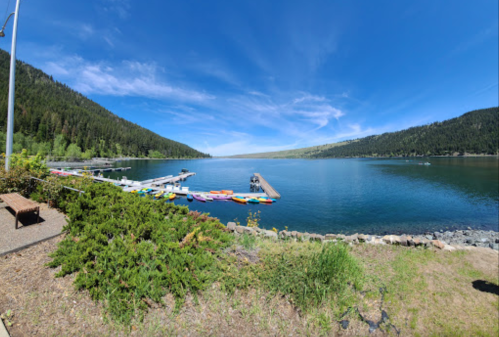 A serene lake surrounded by green hills, with colorful kayaks lined up at a dock under a clear blue sky.