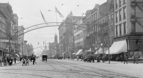 Historic black-and-white street scene with flags, buildings, and early cars, bustling with pedestrians.