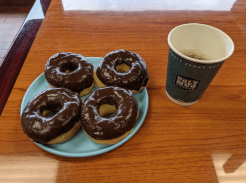 A blue plate with four chocolate-glazed donuts next to a coffee cup on a wooden table.