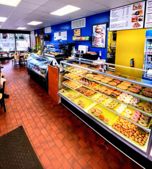 A bright bakery interior featuring a display case filled with various donuts and pastries, with seating in the background.