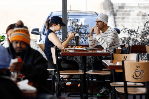 Two people sit at a café table, one using a phone and the other enjoying food, with a car visible outside.