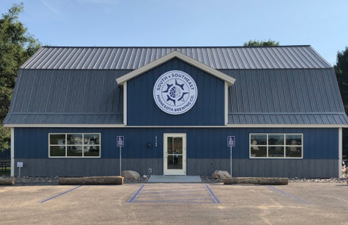 A blue and gray building with a metal roof, featuring a logo and large windows, set in a parking lot.