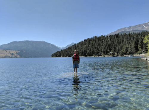 A child stands in shallow water at a lake, surrounded by trees and mountains under a clear blue sky.
