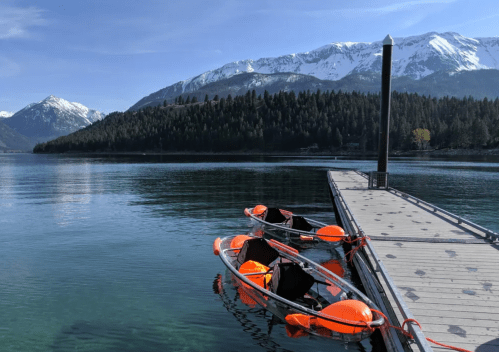 A serene lake scene with a wooden dock and orange kayaks, surrounded by mountains and lush greenery under a clear blue sky.