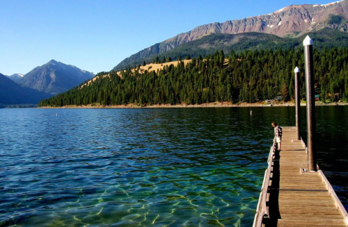 A serene lake with clear water, surrounded by mountains and trees, featuring a wooden pier extending into the water.
