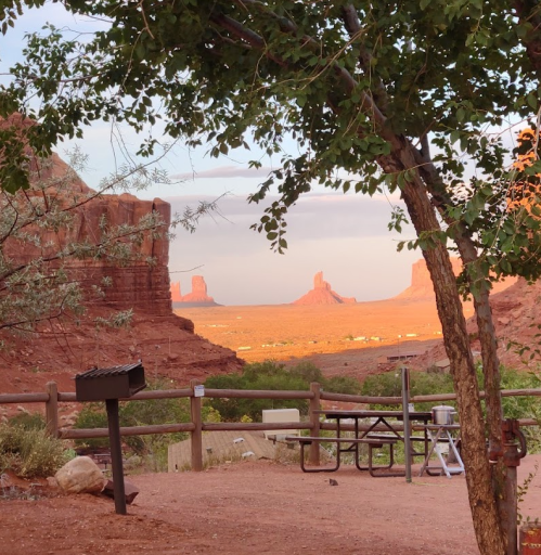 Scenic view of Monument Valley at sunset, framed by trees and a wooden fence, with rock formations in the distance.