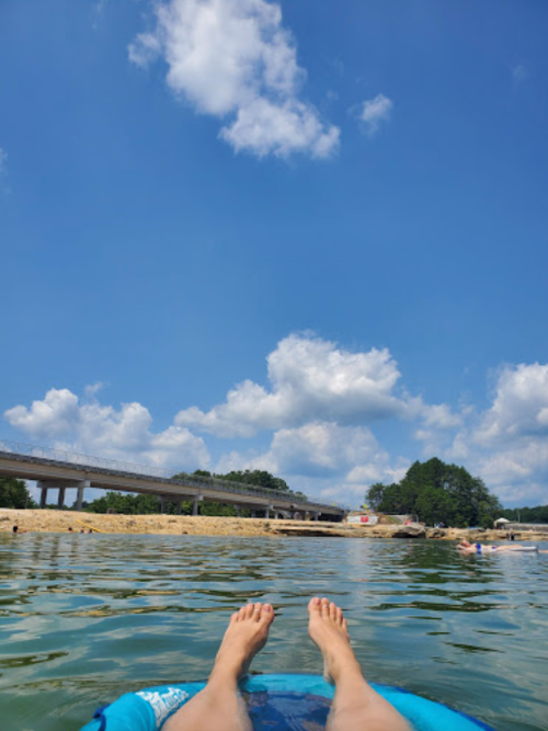 Person's feet on a float in calm water, with a bridge and trees in the background under a blue sky with clouds.