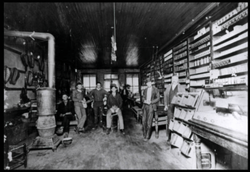 A historic black-and-white photo of men in a shop, surrounded by shelves of goods and a large stove.
